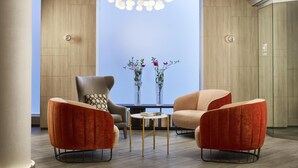 A modern lounge area with a round table, two red upholstered chairs, a grey armchair, and a vase with flowers. - NH Madrid Balboa (Madrid)
