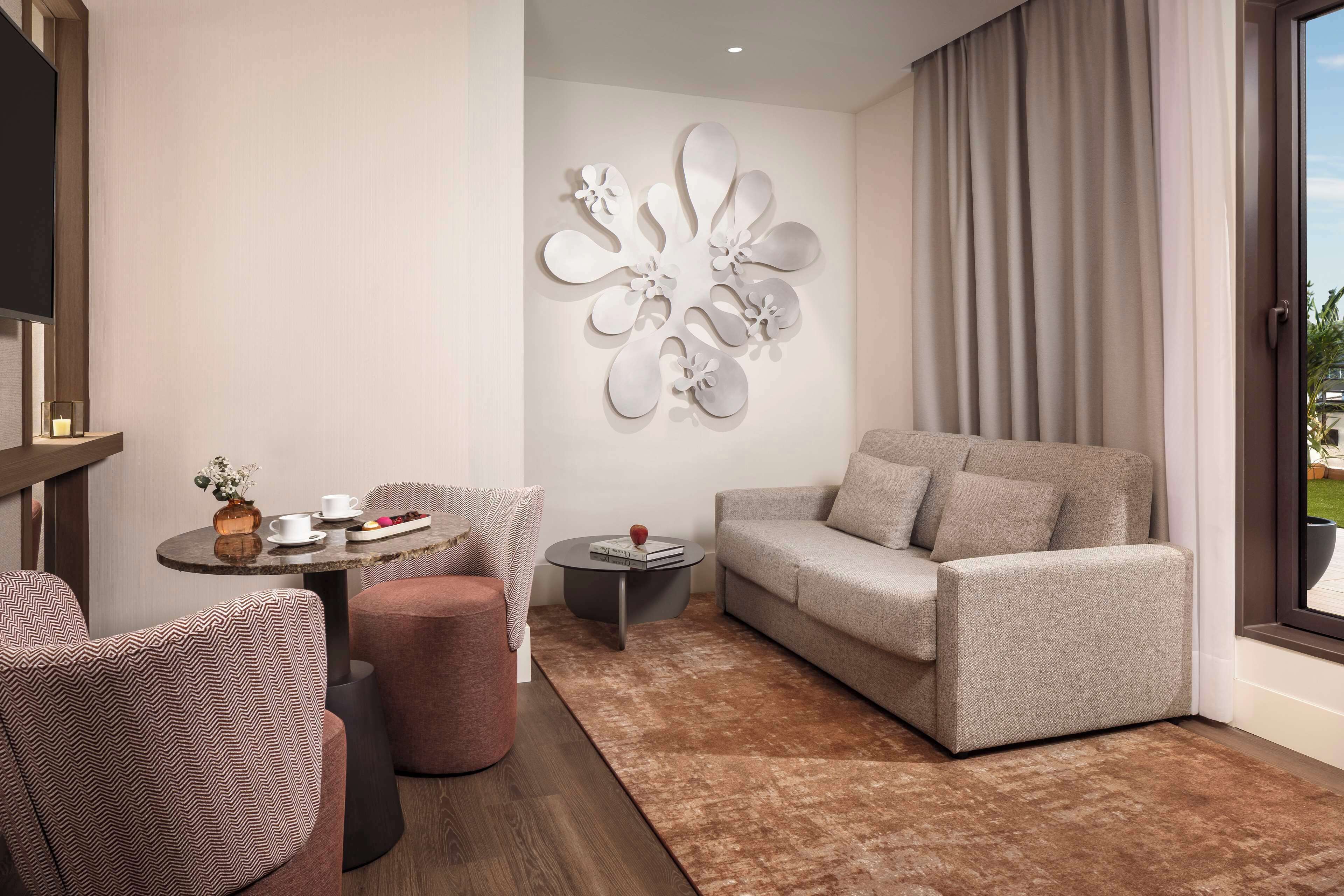A modern living room with a grey sofa, patterned armchairs, a small round table with a vase and books, and a decorative wall piece.