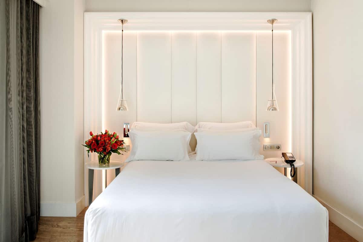 A hotel room with a large bed, two bedside tables, a vase of red flowers, and a telephone on one of the tables.
