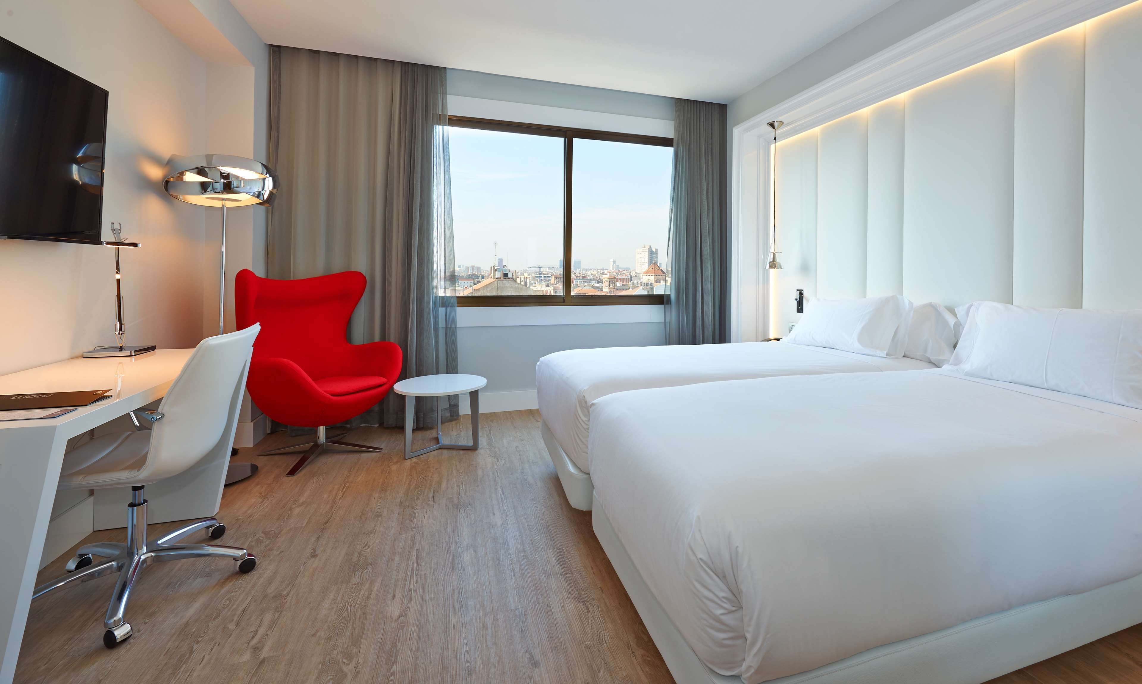 Habitación de hotel con una cama grande, un escritorio con una silla, una silla roja, una mesita redonda pequeña, un televisor montado en la pared y una ventana con vista a la ciudad.