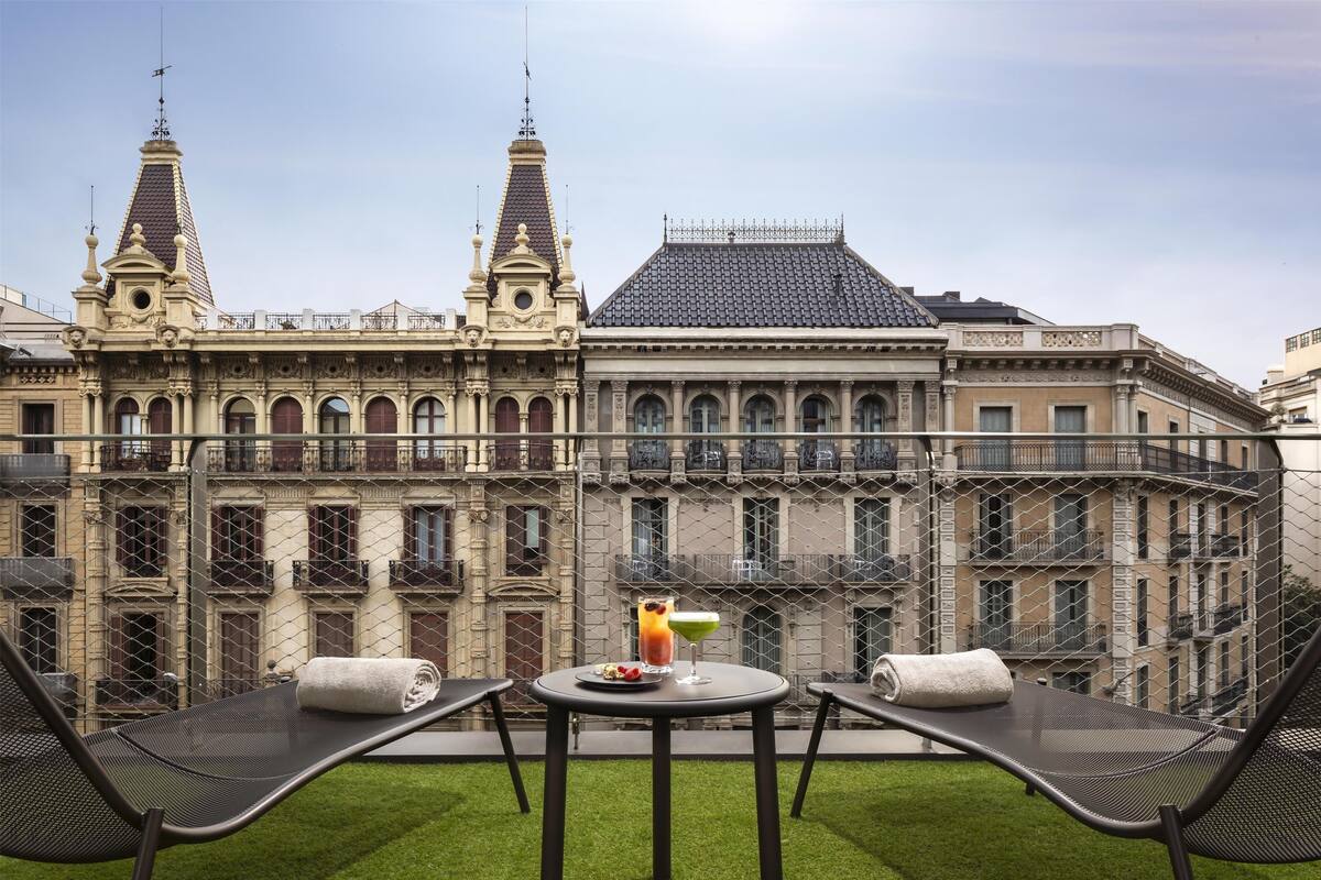 A balcony with two lounge chairs, a small table, and a drink. The background features a historic building with ornate architecture and a tiled roof.