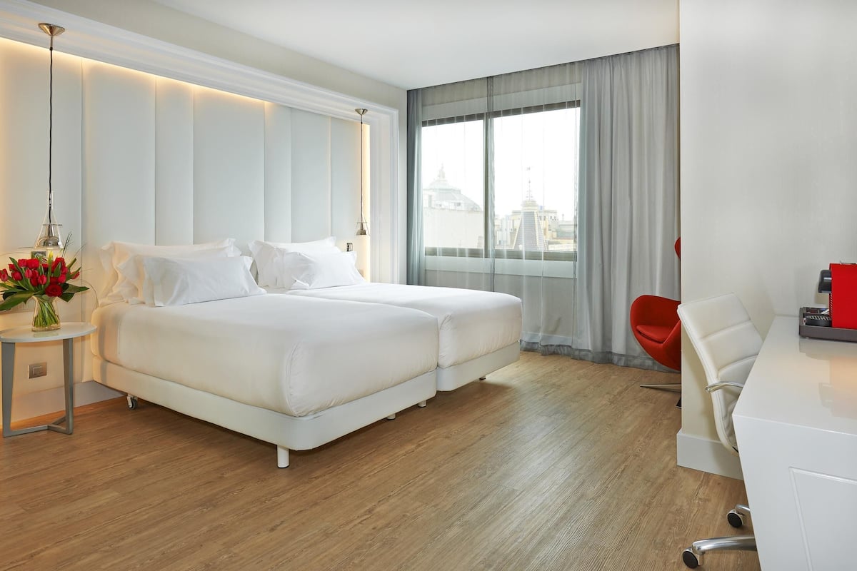 A hotel room with a double bed, a desk, a chair, and a view of a cityscape.