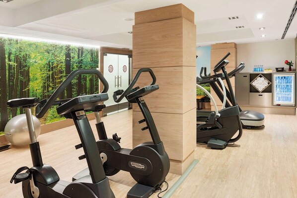 Un gimnasio con bicicletas estáticas y una cinta de correr, suelo de madera y un amplio ventanal con vistas a un bosque.