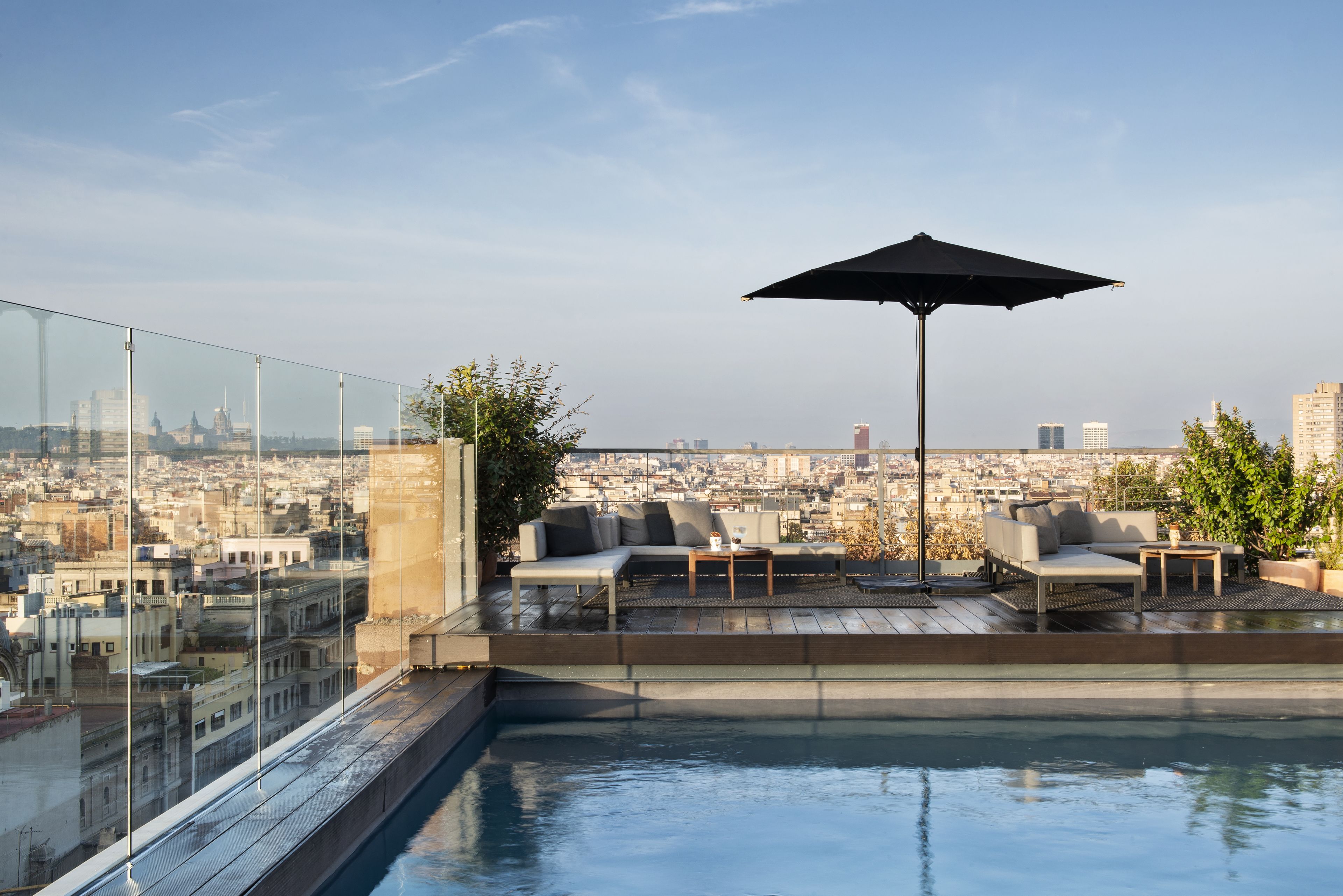Un espace piscine aménagé sur le toit, doté d’une balustrade en verre, d’un coin salon avec un canapé et des fauteuils, et d’un grand parasol.