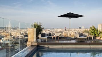Un espace piscine aménagé sur le toit, doté d’une balustrade en verre, d’un coin salon avec un canapé et des fauteuils, et d’un grand parasol.