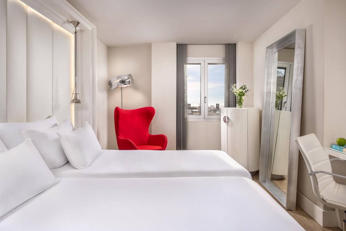 A hotel room with a large bed, a red chair, a desk with a chair, a mirror, and a window with a view of the city.