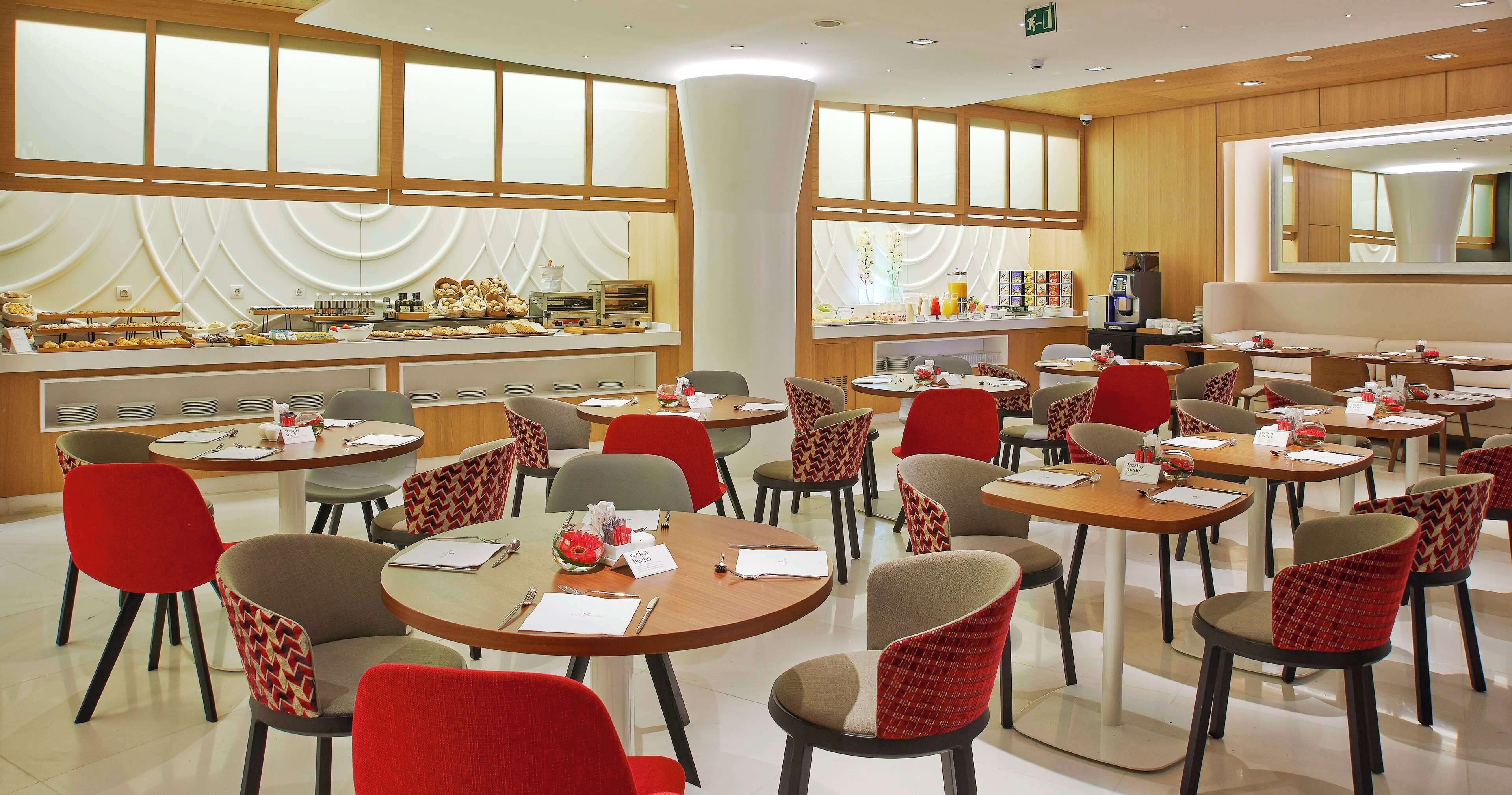 A hotel dining area with round tables, red and grey chairs, a buffet with pastries, and a coffee machine.