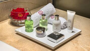 A bathroom vanity with a marble countertop, a white towel, a red flower in a glass bowl, and various toiletries including a shower gel, shampoo, body milk, hand cream, and a soft mask.