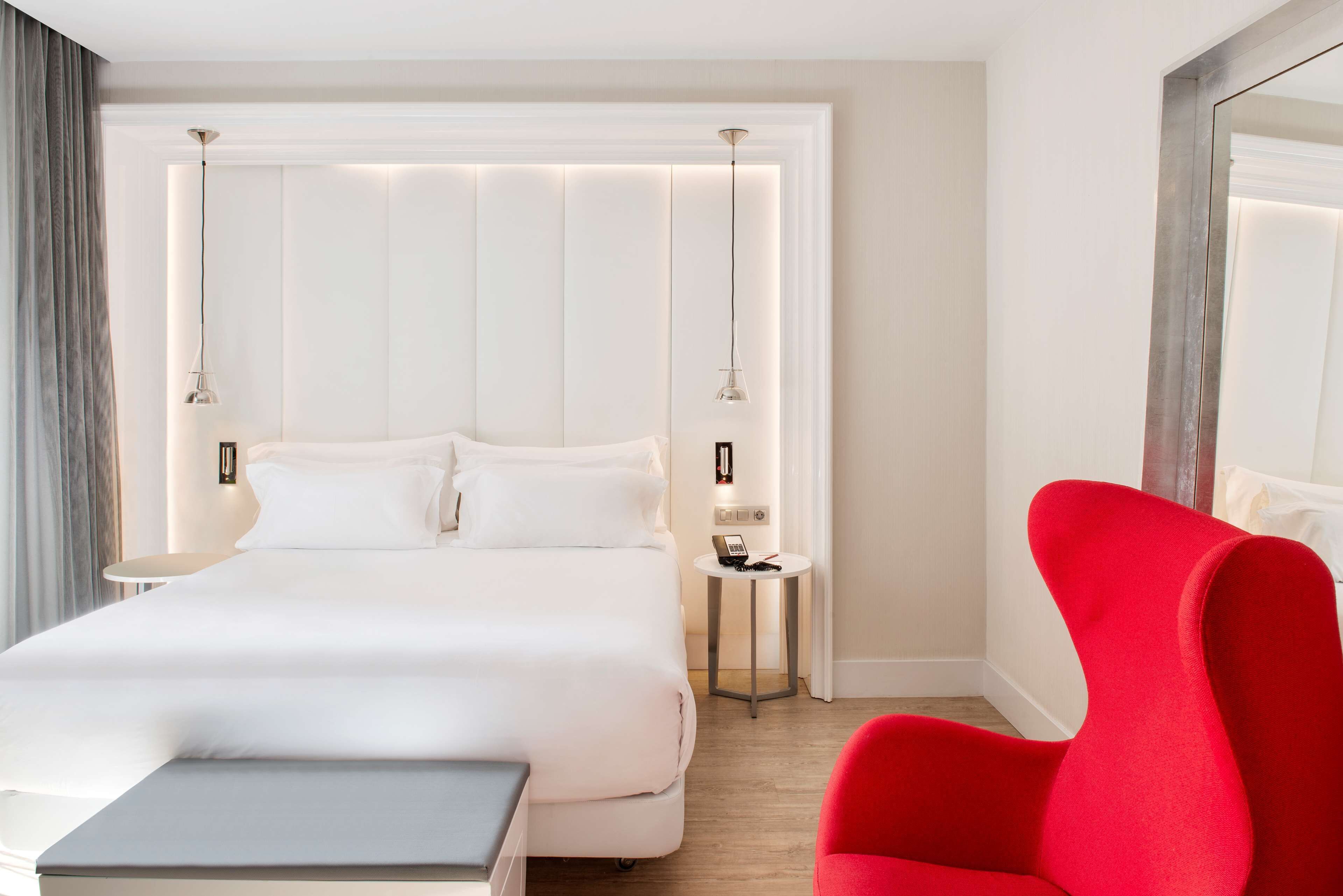 A modern hotel room with a large bed, bedside tables, a red chair, and a small table.