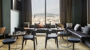 Quarto moderno com sofá, cadeiras e mesas dispostas em torno de uma grande janela com vista para a cidade.