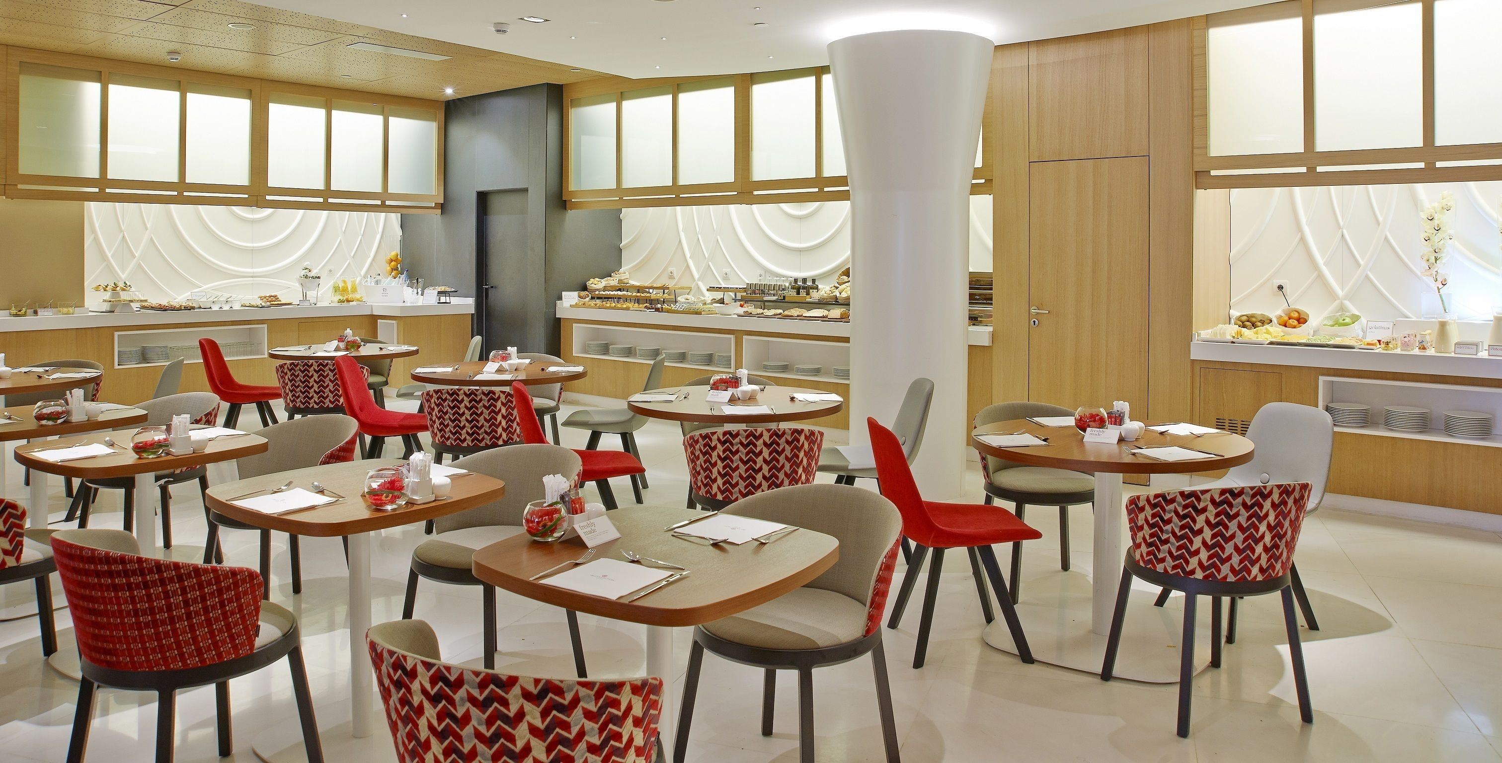 A modern dining area with multiple tables and chairs, a large pillar, and a buffet counter with various food items.