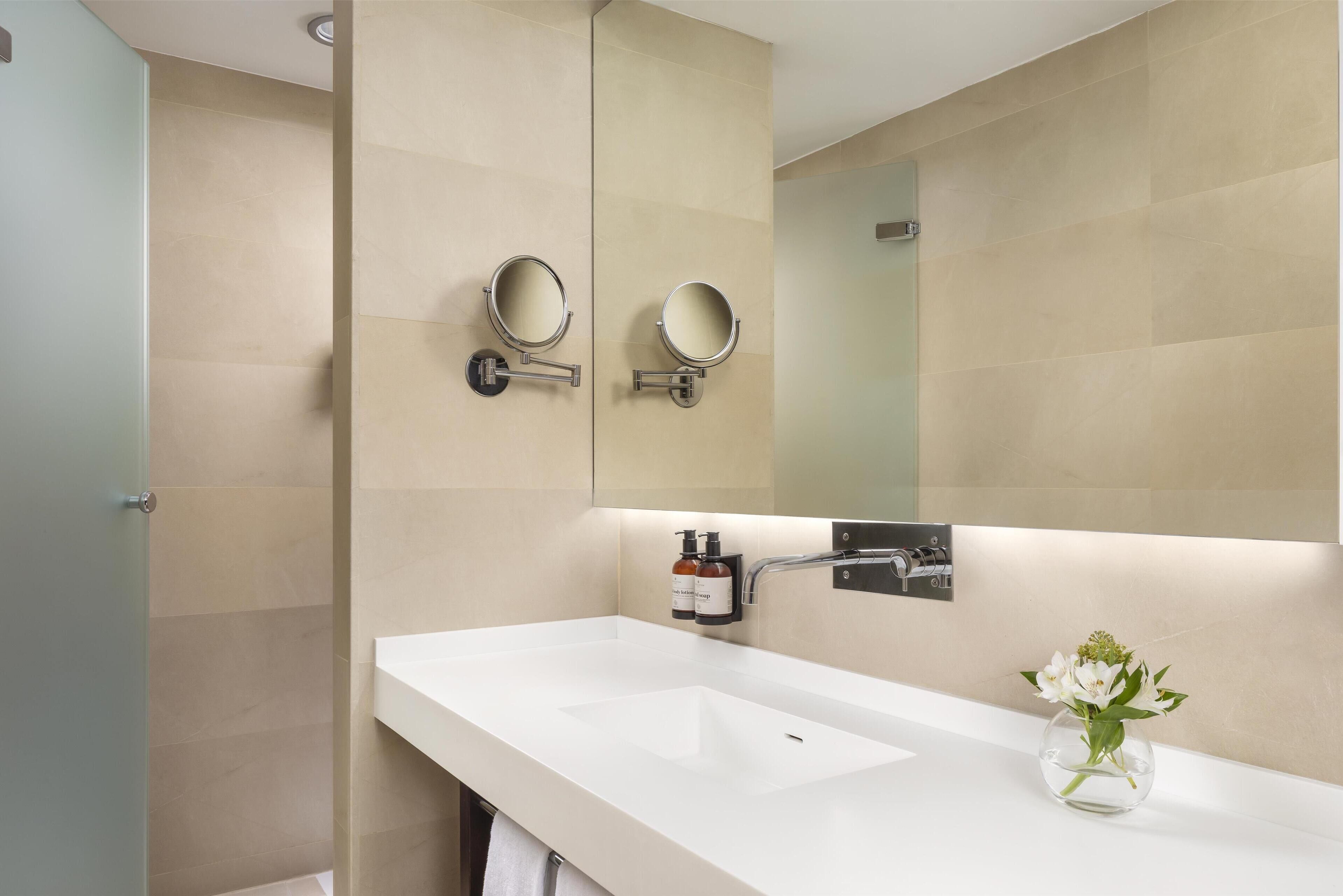 Un baño moderno con un lavabo blanco, dos espejos adosados a la pared, un perchero y un jarrón con flores.