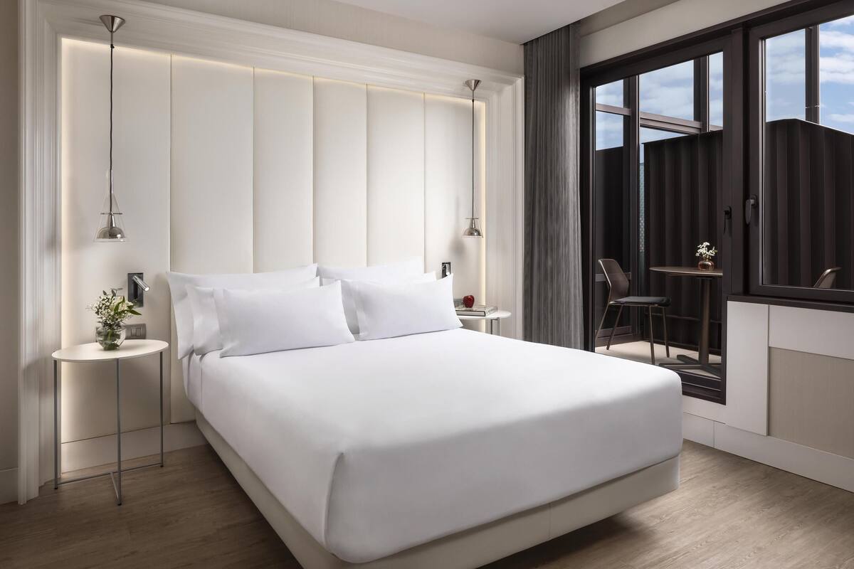 A modern hotel room with a large bed, bedside tables, a small table with a vase, and a view of the outdoors through large windows.