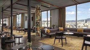 Um hall de hotel moderno com área de bar, espaços de estar e uma grande janela com vista para a cidade.