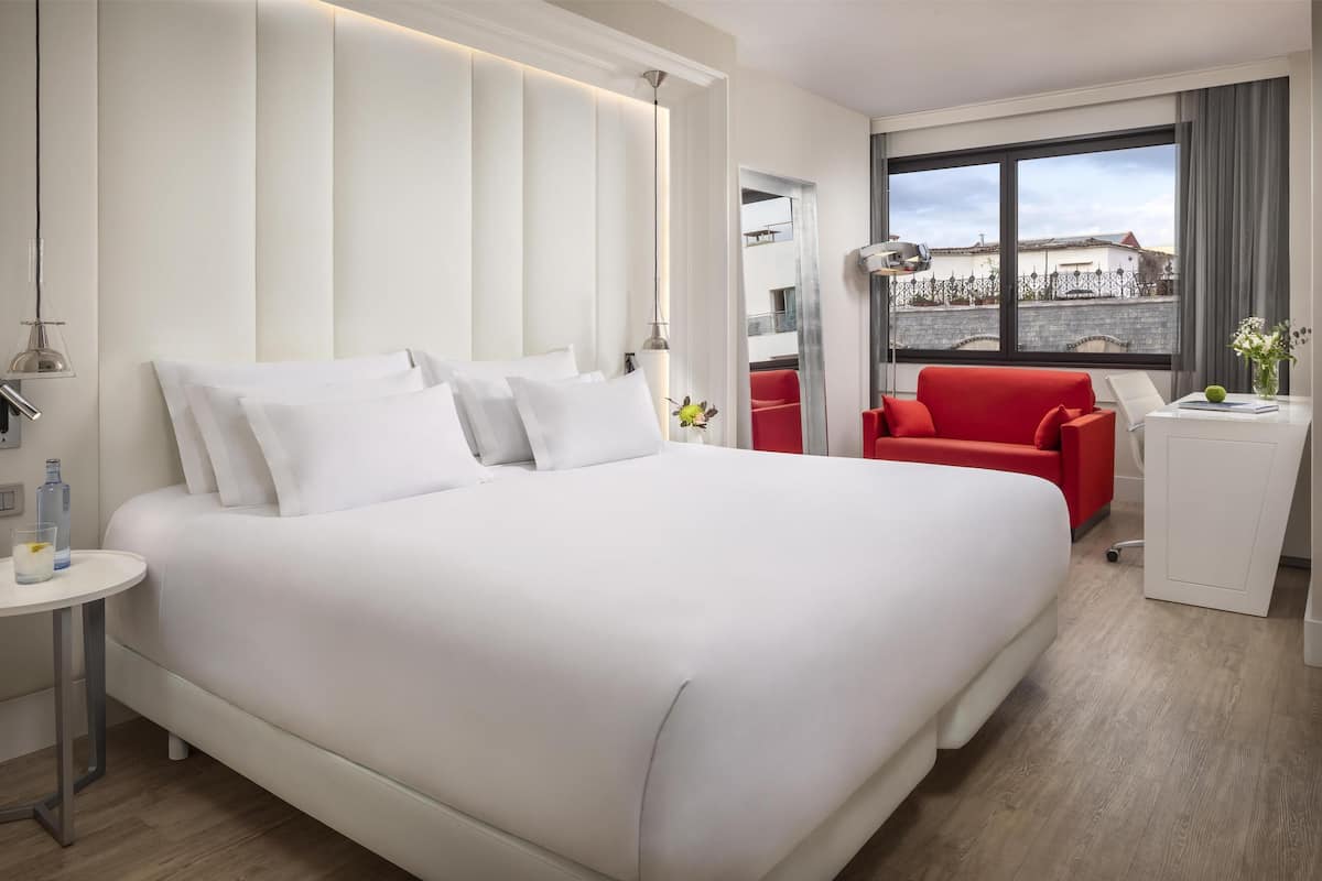 A modern hotel room with a large bed, a red armchair, a small desk, and a view of the city through large windows.