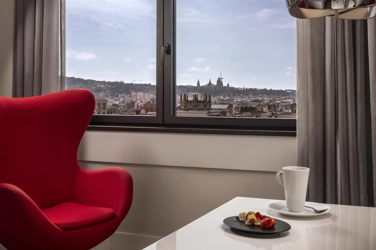 A modern room with a red chair, a white table with a plate of food and a cup, and a window with a city view.