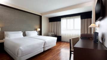Habitación de hotel con dos camas, un escritorio, una silla, un televisor y un ventanal grande con cortinas.