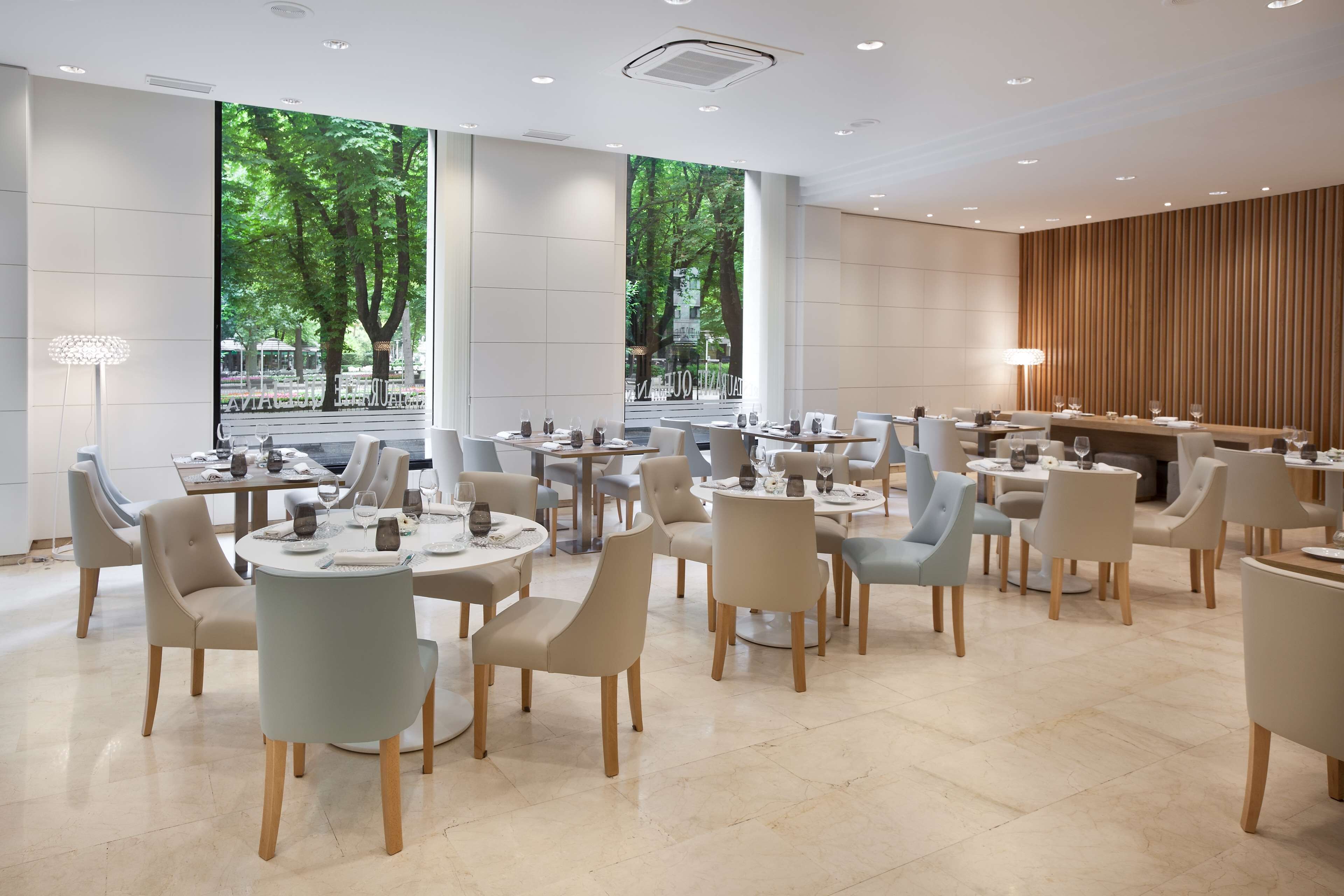 A modern dining area with round tables and chairs, large windows with a view of trees, and a wooden accent wall.