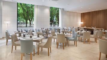 A modern dining area with round tables and chairs, large windows with a view of trees, and a wooden accent wall.