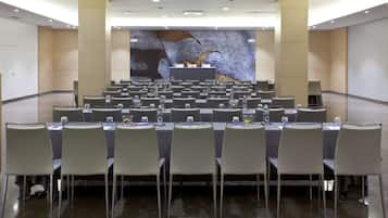 Un salón de conferencias con varias filas de sillas y mesas, un atril y un gran mural en la pared de fondo.