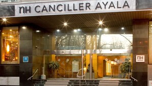 La entrada al hotel NH Canciller Ayala, con una fachada moderna, puertas de cristal y una escalera que conduce a la recepción.