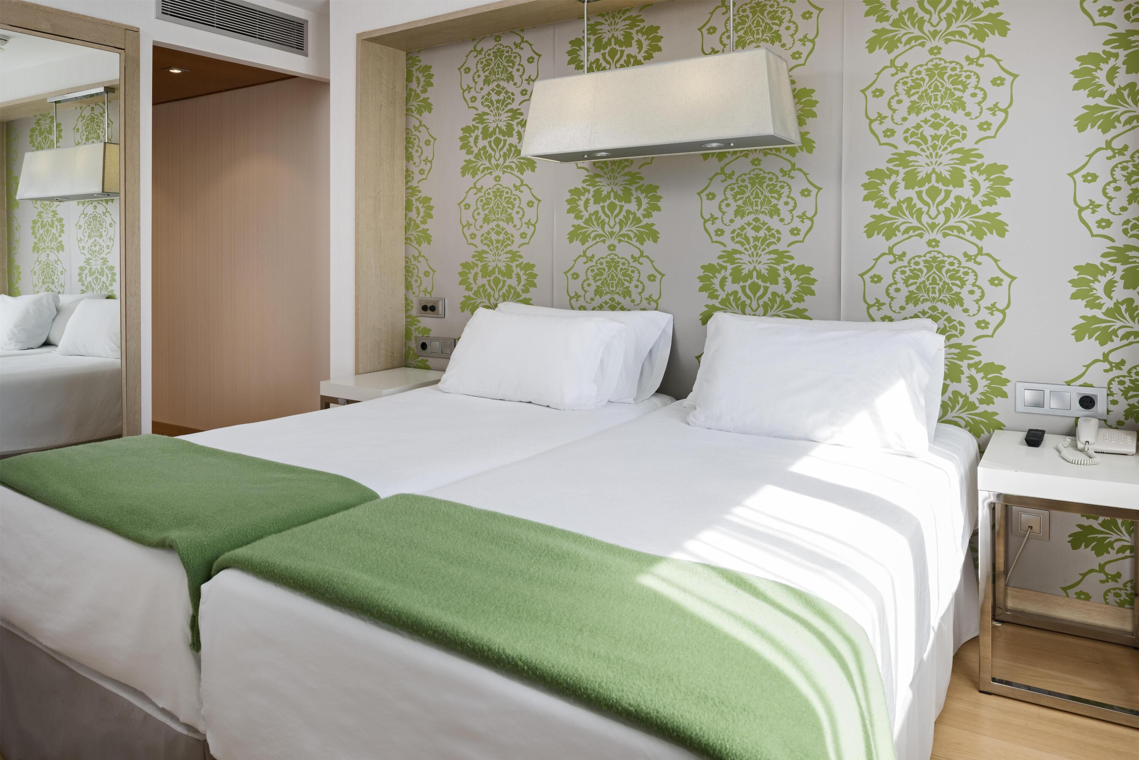 A hotel room with two beds, a bedside table with a phone, and a mirror.