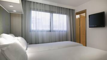 A hotel room with a bed, a television on the wall, and a window with curtains.