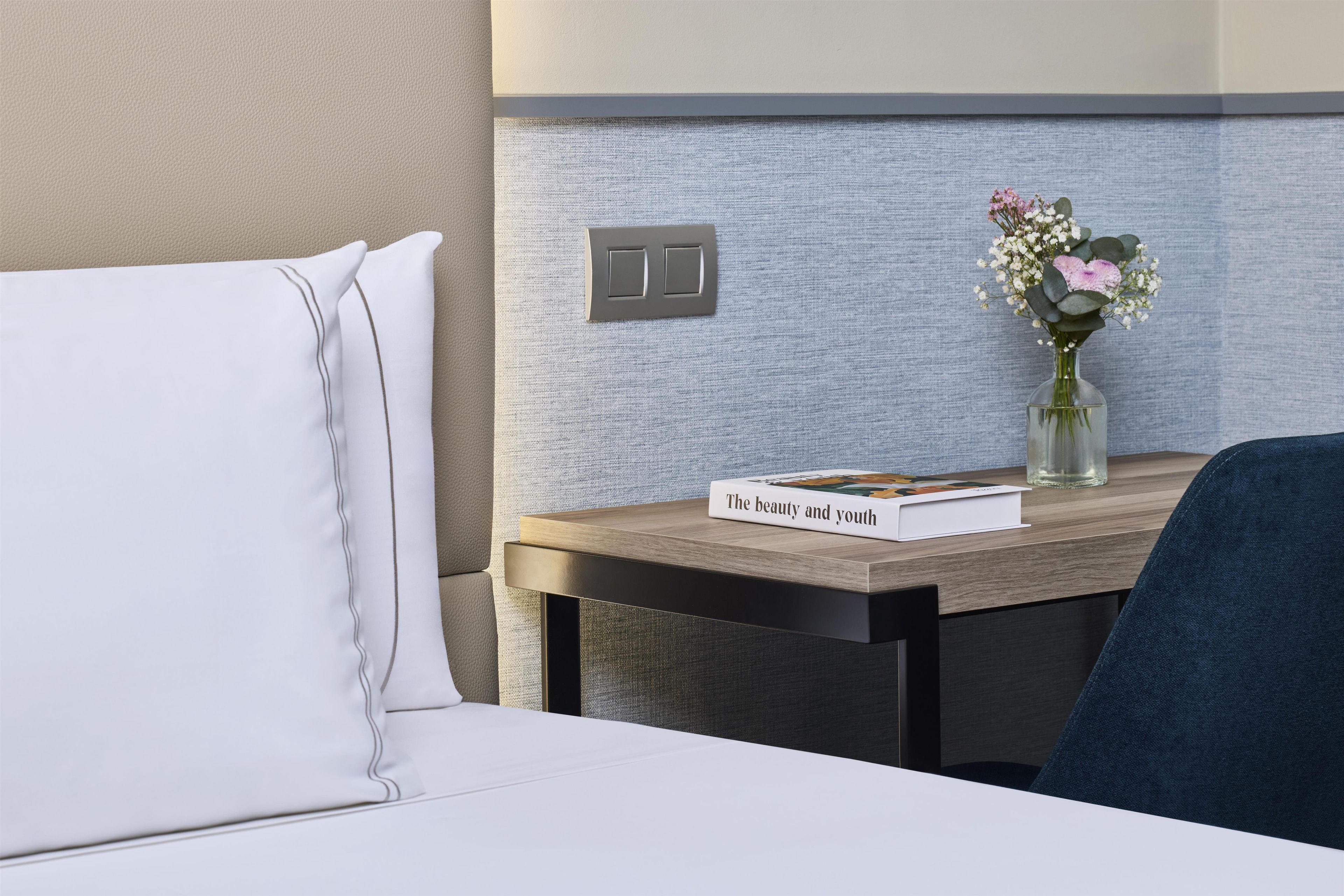 A hotel room with a bed, a bedside table with a book and a vase of flowers, and a wall-mounted light switch.