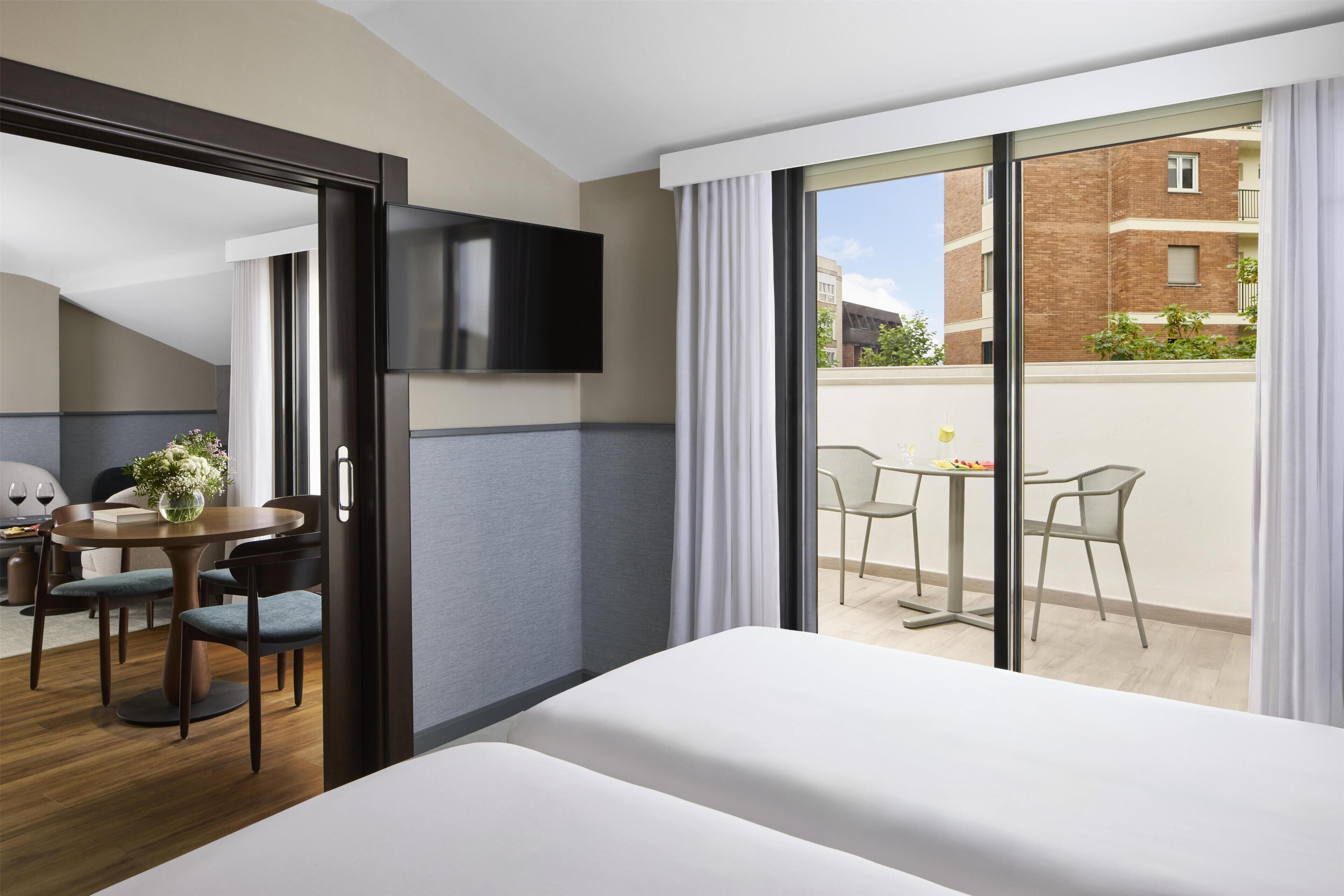 Une chambre d’hôtel comprenant un lit, une télévision, un coin repas avec une table et des chaises, ainsi qu’un balcon offrant une vue sur les bâtiments.