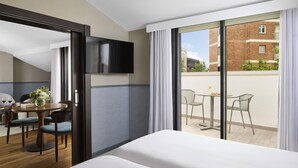 A hotel room with a bed, a TV, a dining area with a table and chairs, and a balcony with a view of buildings. - NH Ciudad de Santander (Santander)