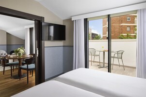 Habitación de hotel con cama, televisión, zona de comedor con mesa y sillas, y balcón con vistas a edificios.