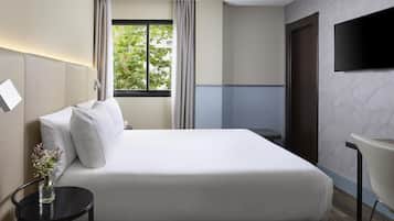 A modern hotel room with a large bed, a bedside table with a vase of flowers, a flat-screen TV mounted on the wall, and a window with curtains.