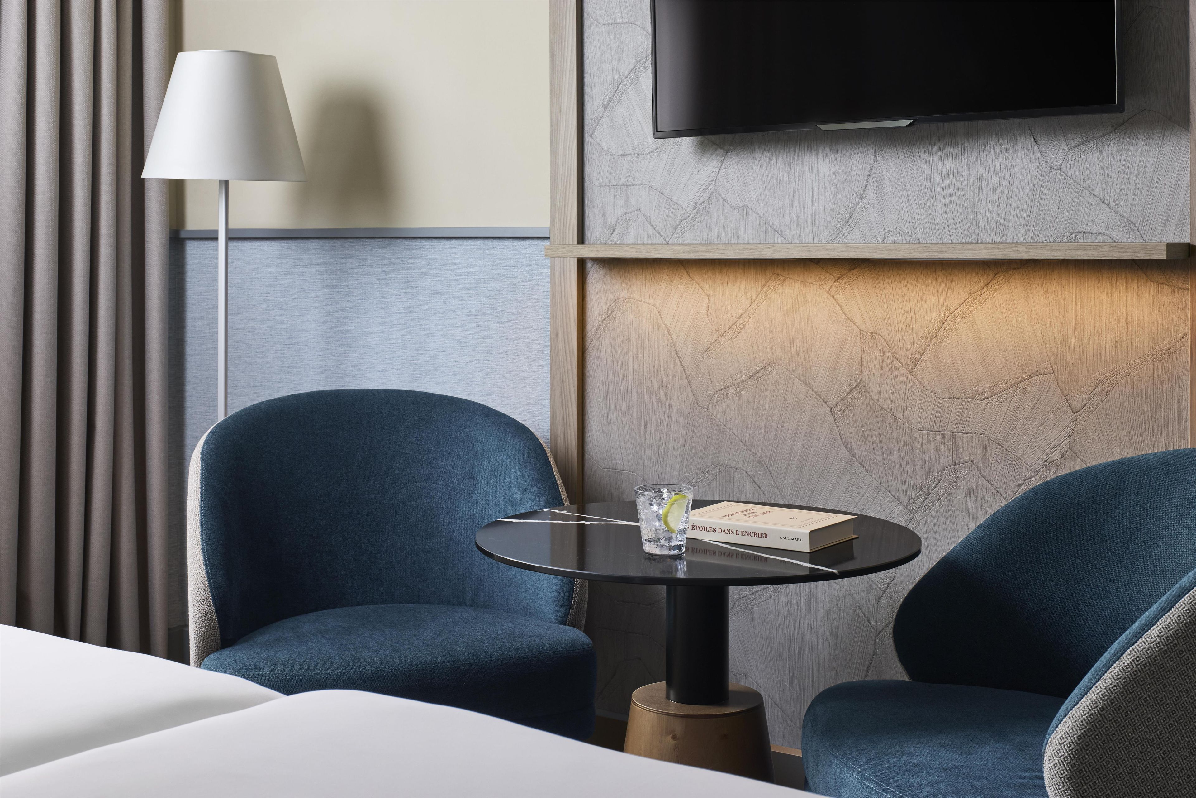 Une chambre d’hôtel avec un lit, deux fauteuils bleus, une petite table ronde sur laquelle se trouvent un livre et un verre d’eau, une lampe et un téléviseur fixé au mur.