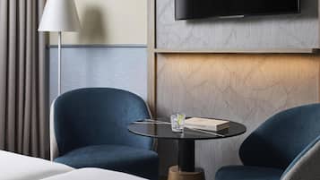 Une chambre d’hôtel avec un lit, deux fauteuils bleus, une petite table ronde sur laquelle se trouvent un livre et un verre d’eau, une lampe et un téléviseur fixé au mur.