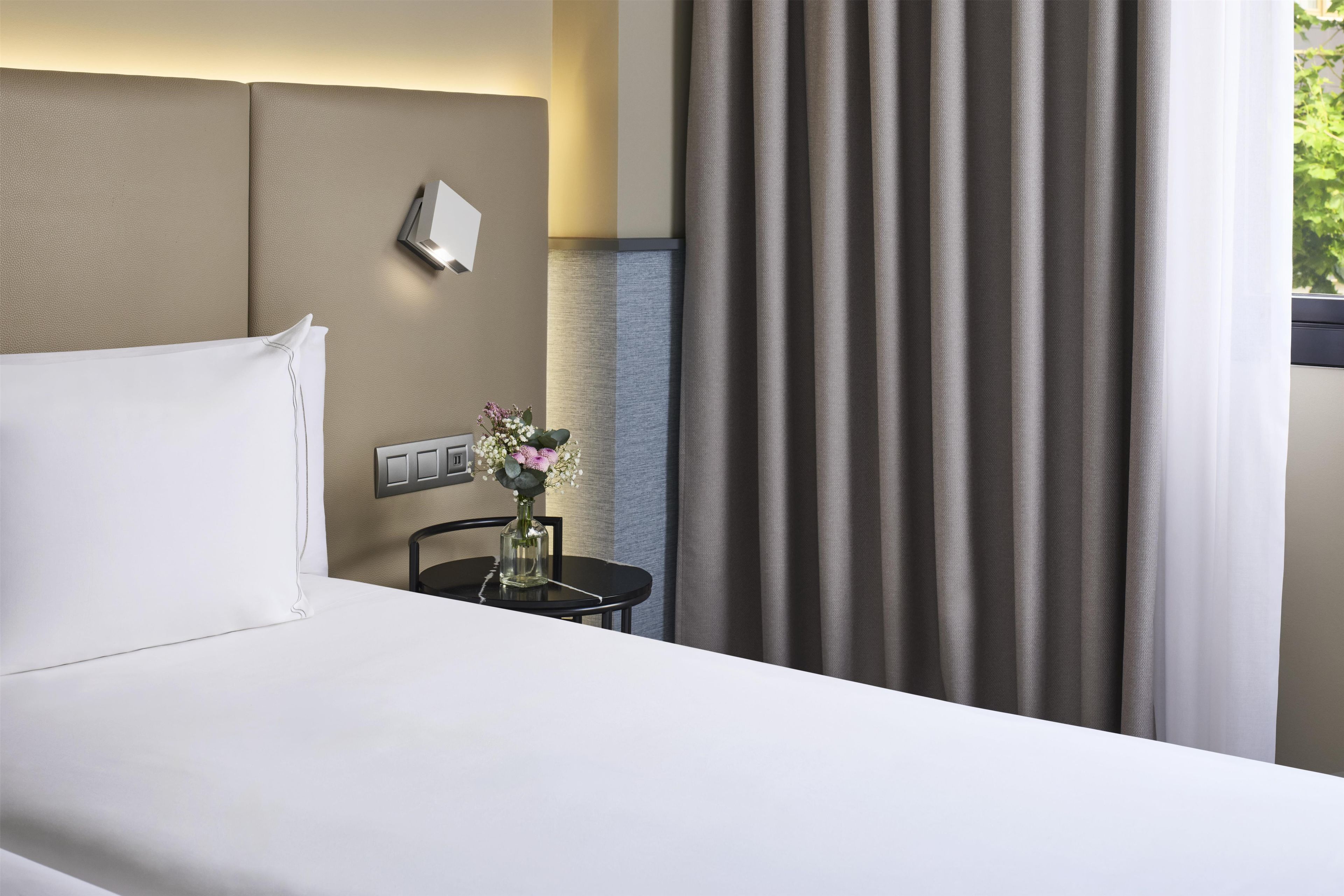 A hotel room with a bed, a nightstand, a vase with flowers, a wall-mounted light, and a window with curtains.