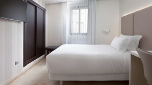 Una habitación de hotel moderna con una cama grande, un armario oscuro, una mesita pequeña y una ventana con cortinas traslúcidas.