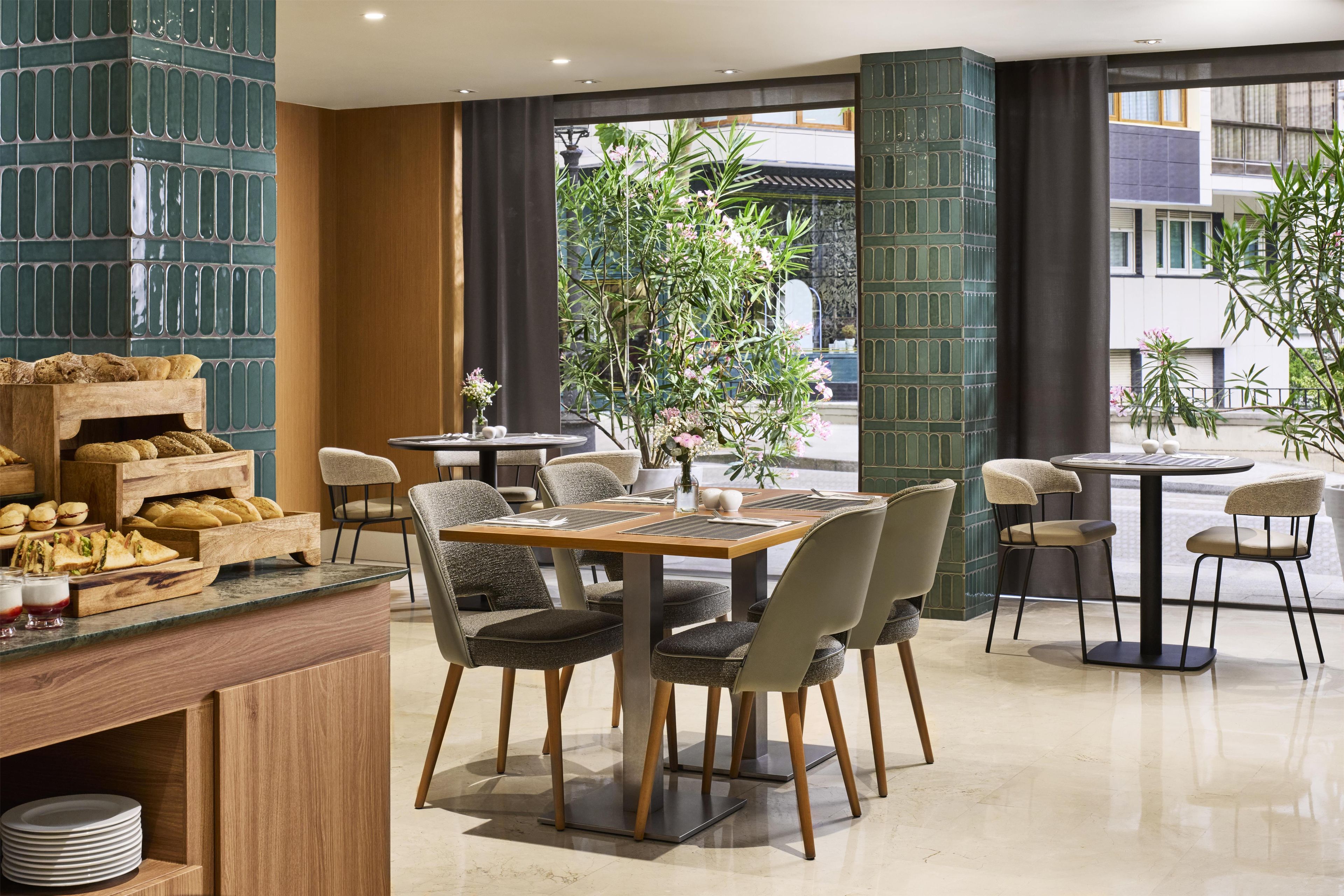 Een modern restaurantinterieur met een centrale eettafel, stoelen en een buffet met brood en gebak.