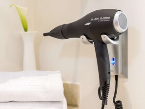 A black hairdryer mounted on a wall, a white towel, and a vase with a green plant. - NH Ciudad de Santander (Santander)