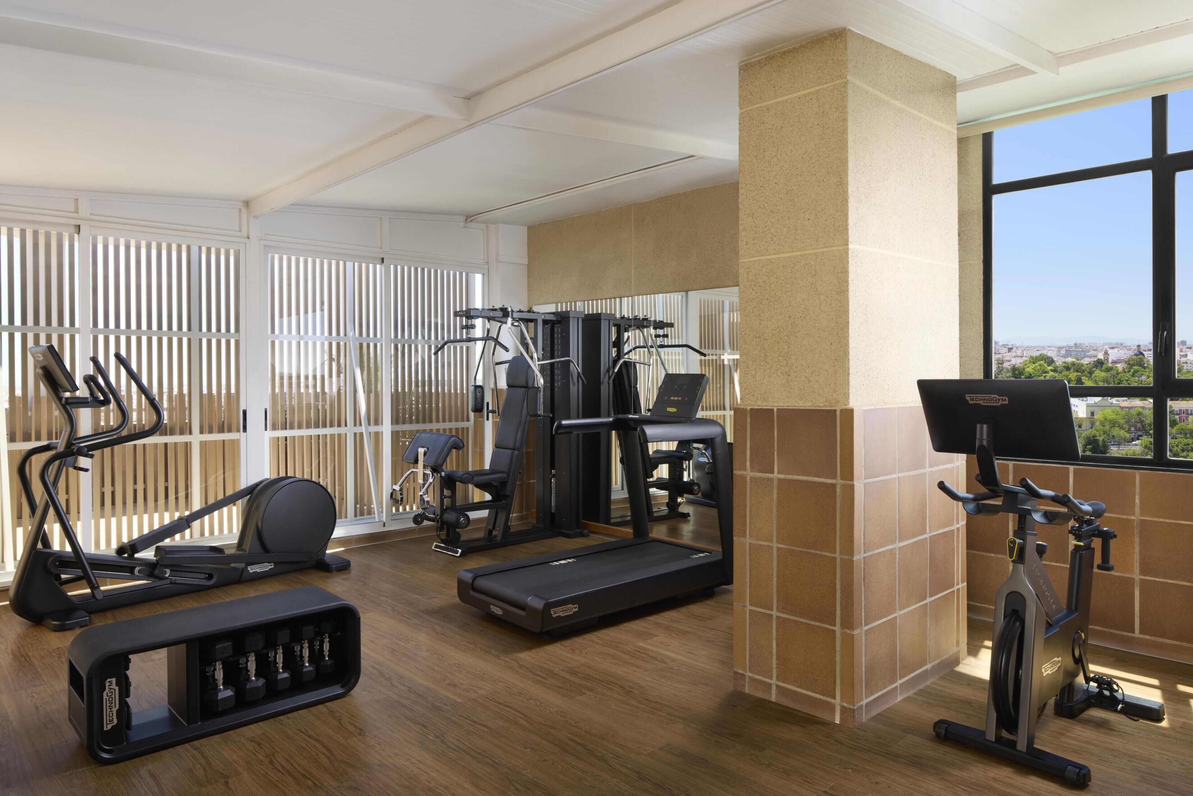 Un gimnasio con cintas de correr, elípticas y máquinas de pesas, ventanas amplias con vista a la ciudad y piso de madera.