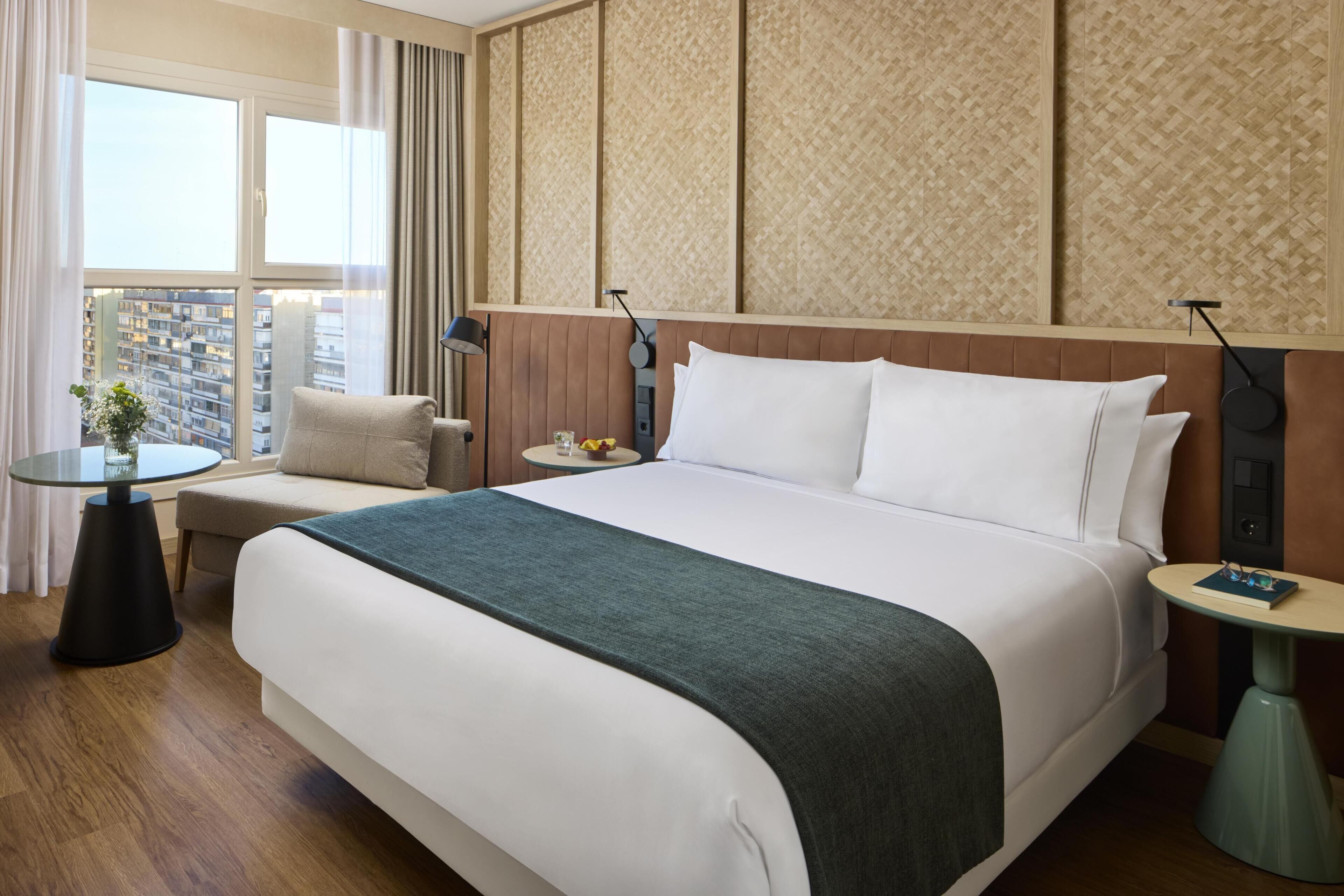 A hotel room with a large bed, a small round table, a chair, a nightstand, and a view of the city.