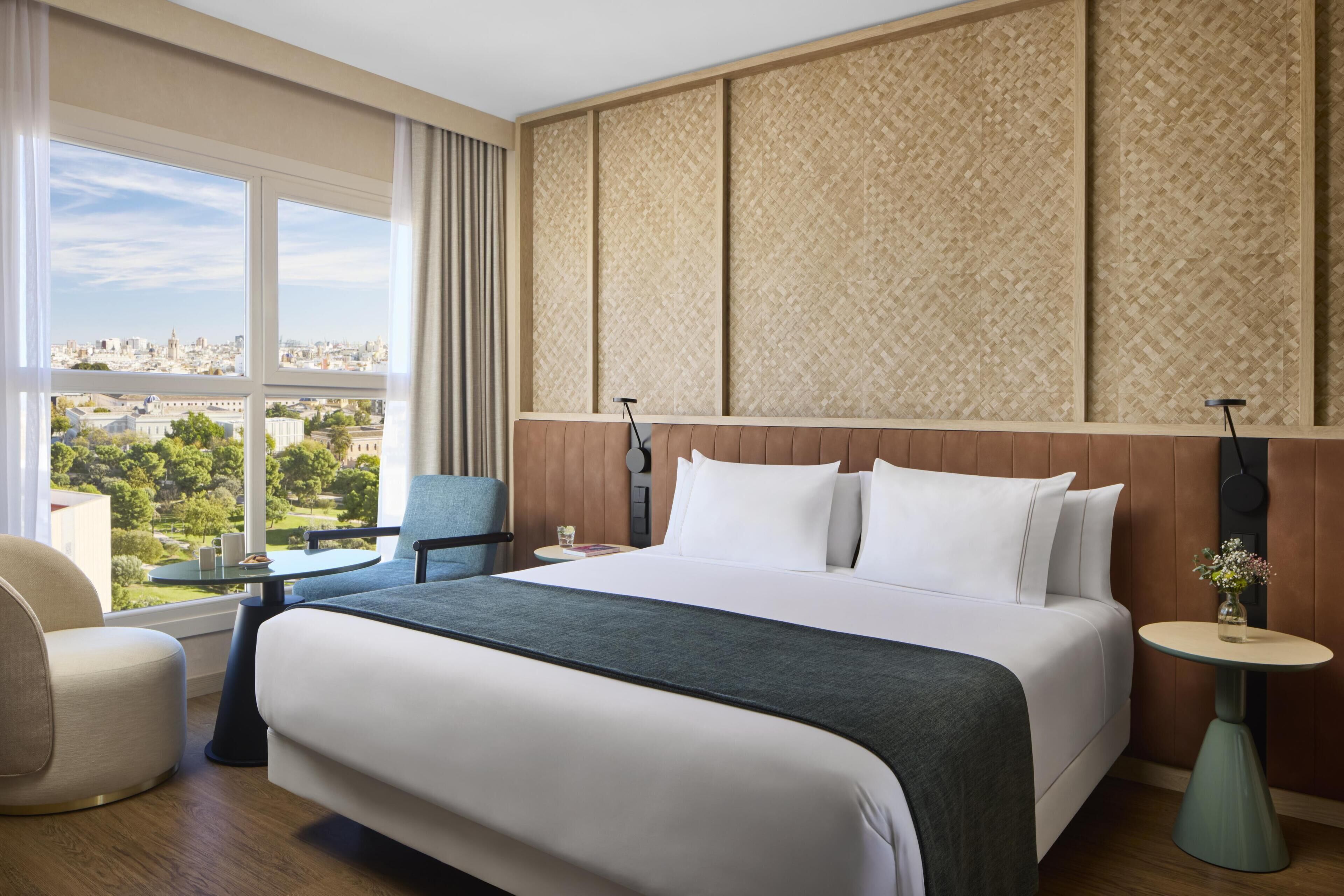 Een hotelkamer met een groot bed, uitzicht op de stad, een klein tafeltje, een stoel en een decoratief wandpaneel.
