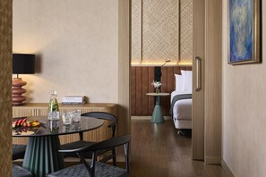 A hotel room with a dining table, chairs, a sofa, a small side table, a lamp, a painting, and a bed. - NH Valencia Center (Valencia)