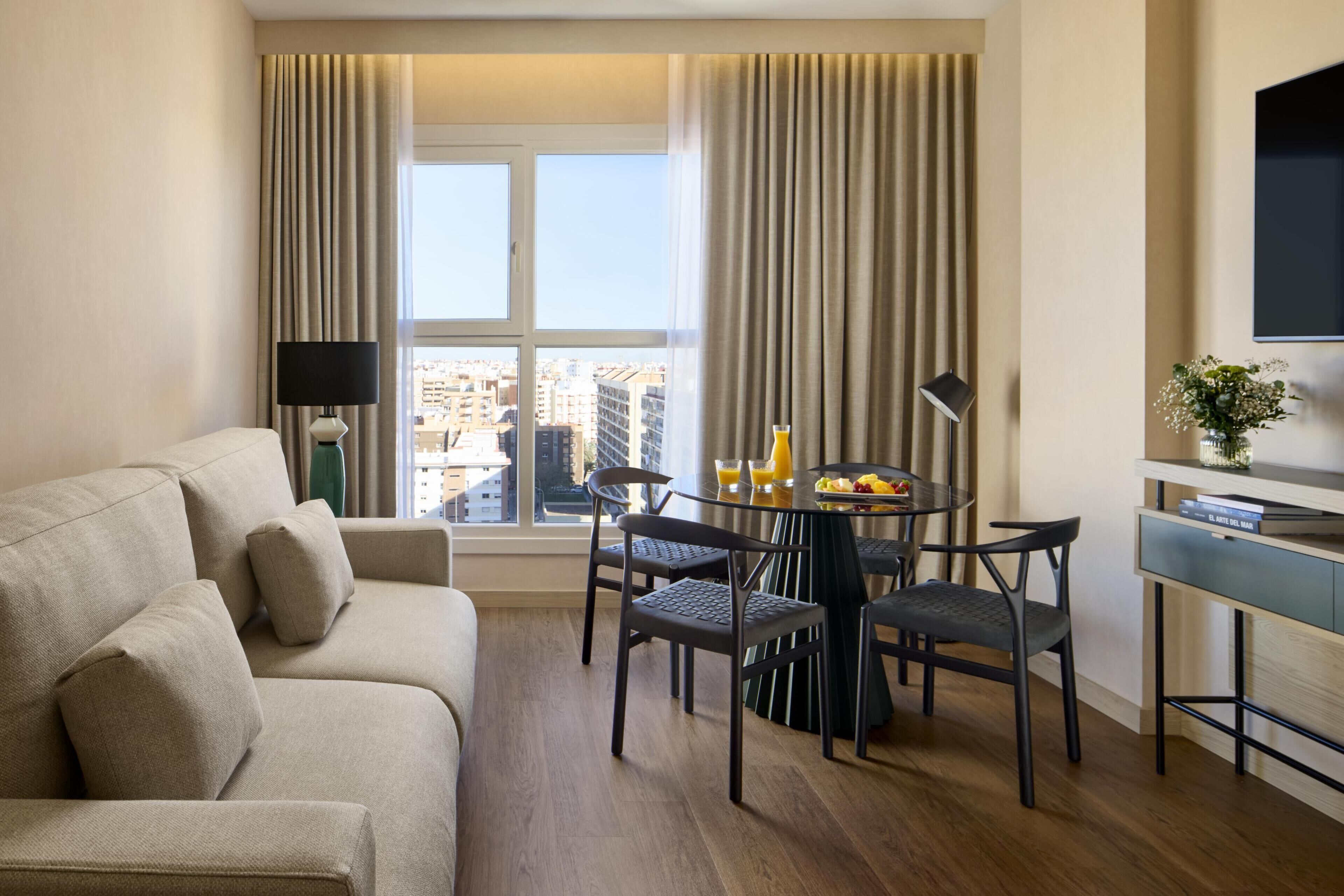 Una habitación de hotel moderna con sofá, mesa de comedor, sillas, televisor y vistas a la ciudad.