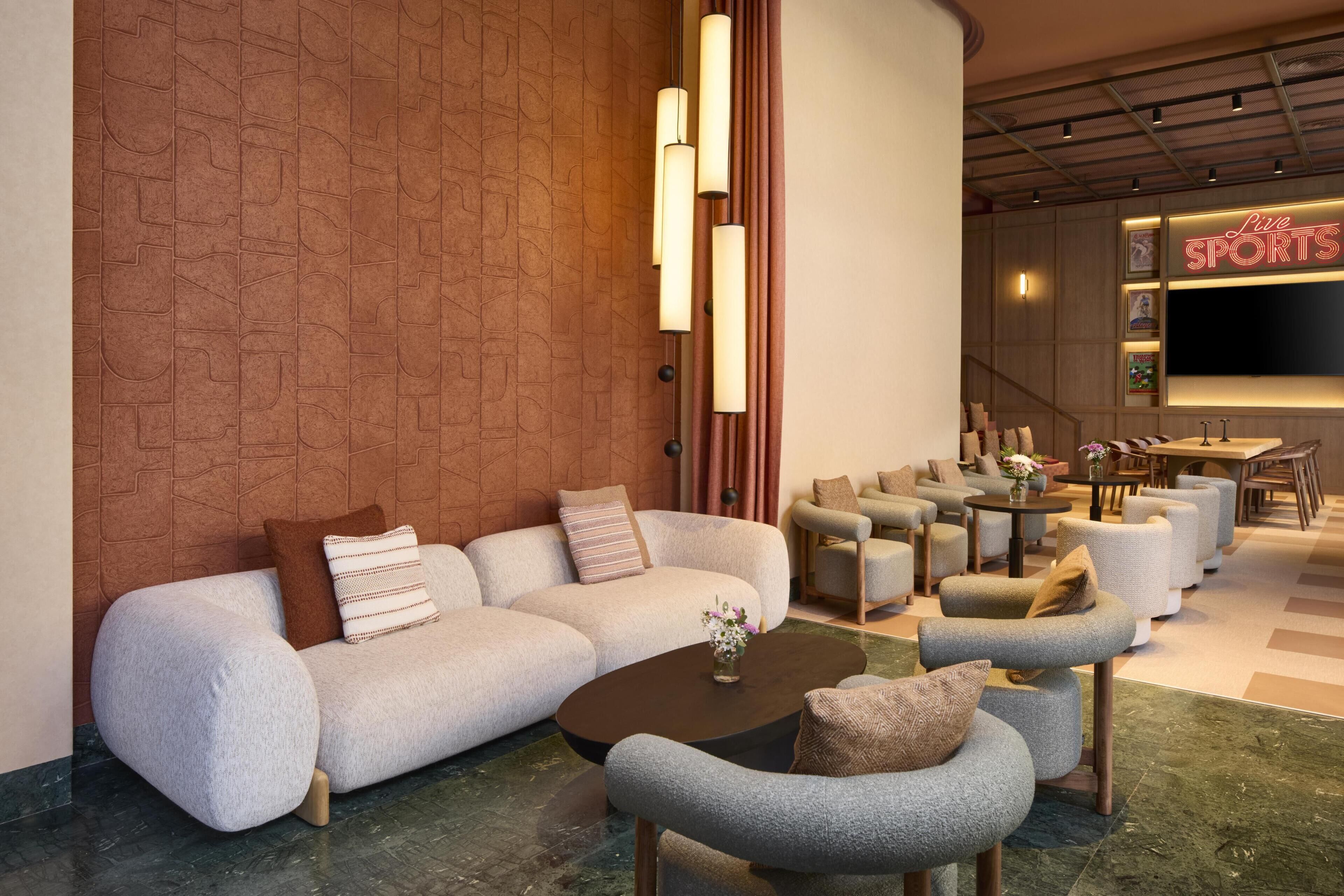A modern hotel lobby with a sectional sofa, armchairs, a coffee table, a TV screen, and a staircase.