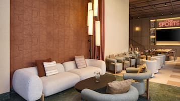 A modern hotel lobby with a sectional sofa, armchairs, a coffee table, a TV screen, and a staircase.
