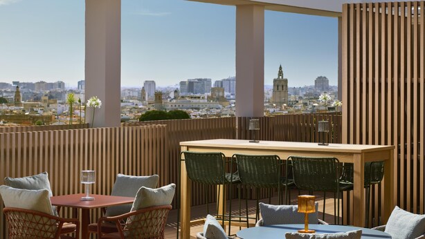 Una terraza en la azotea con vistas a la ciudad, que cuenta con una mesa de madera, sillas de mimbre y una lámpara pequeña.