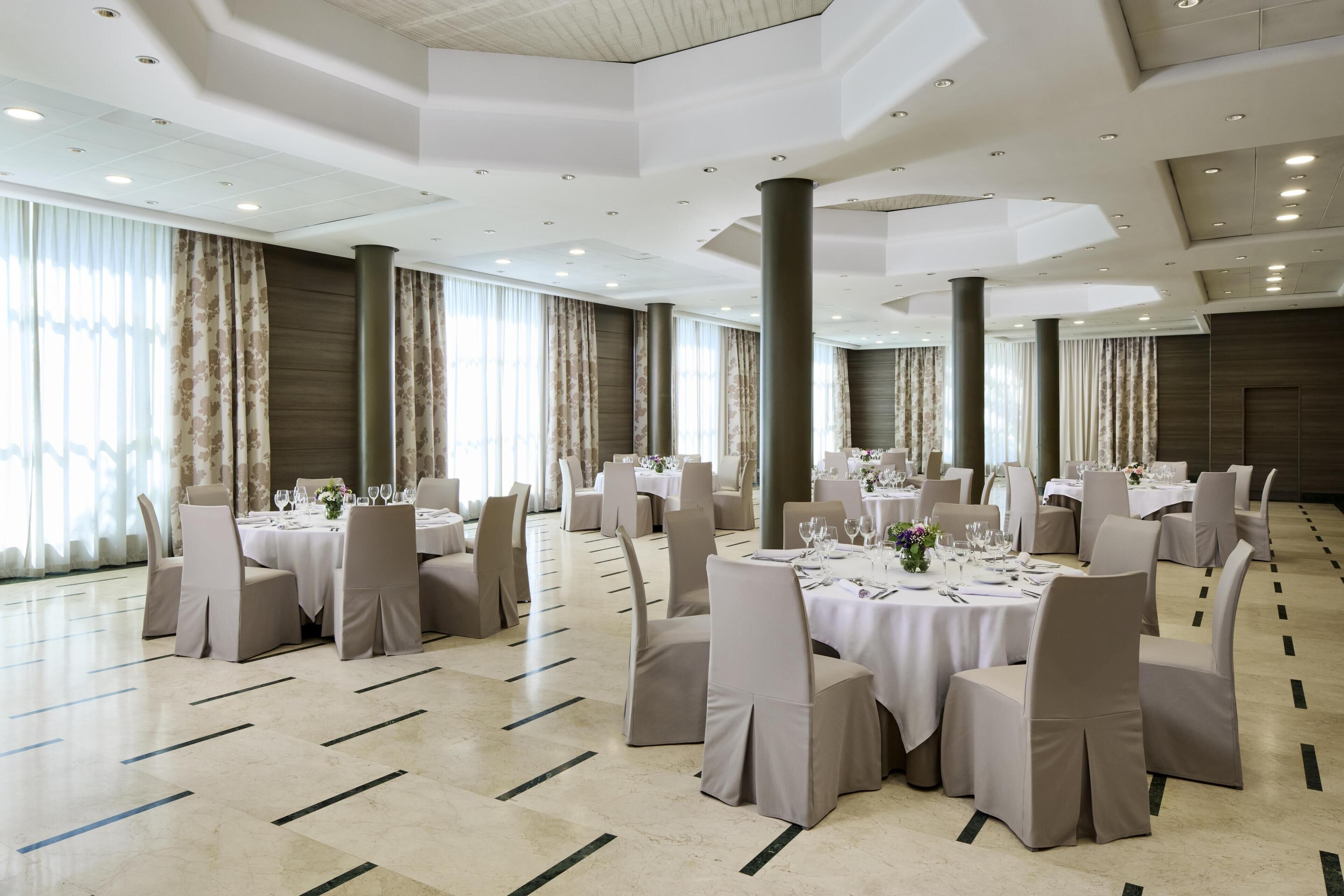 A spacious banquet hall with round tables, beige chairs, and a high ceiling with recessed lighting.