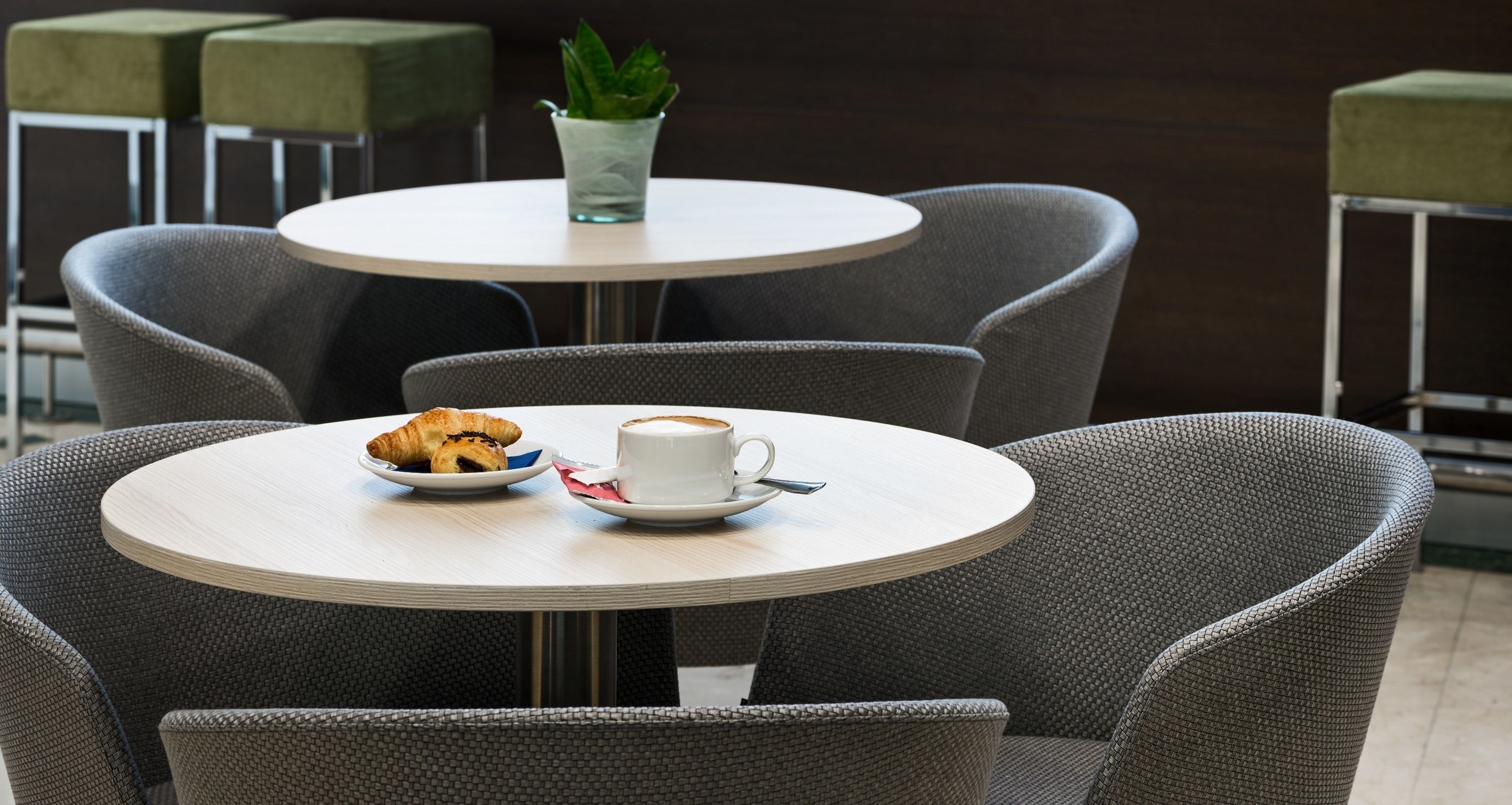 A round table with a croissant and a cup of coffee, surrounded by grey upholstered chairs.