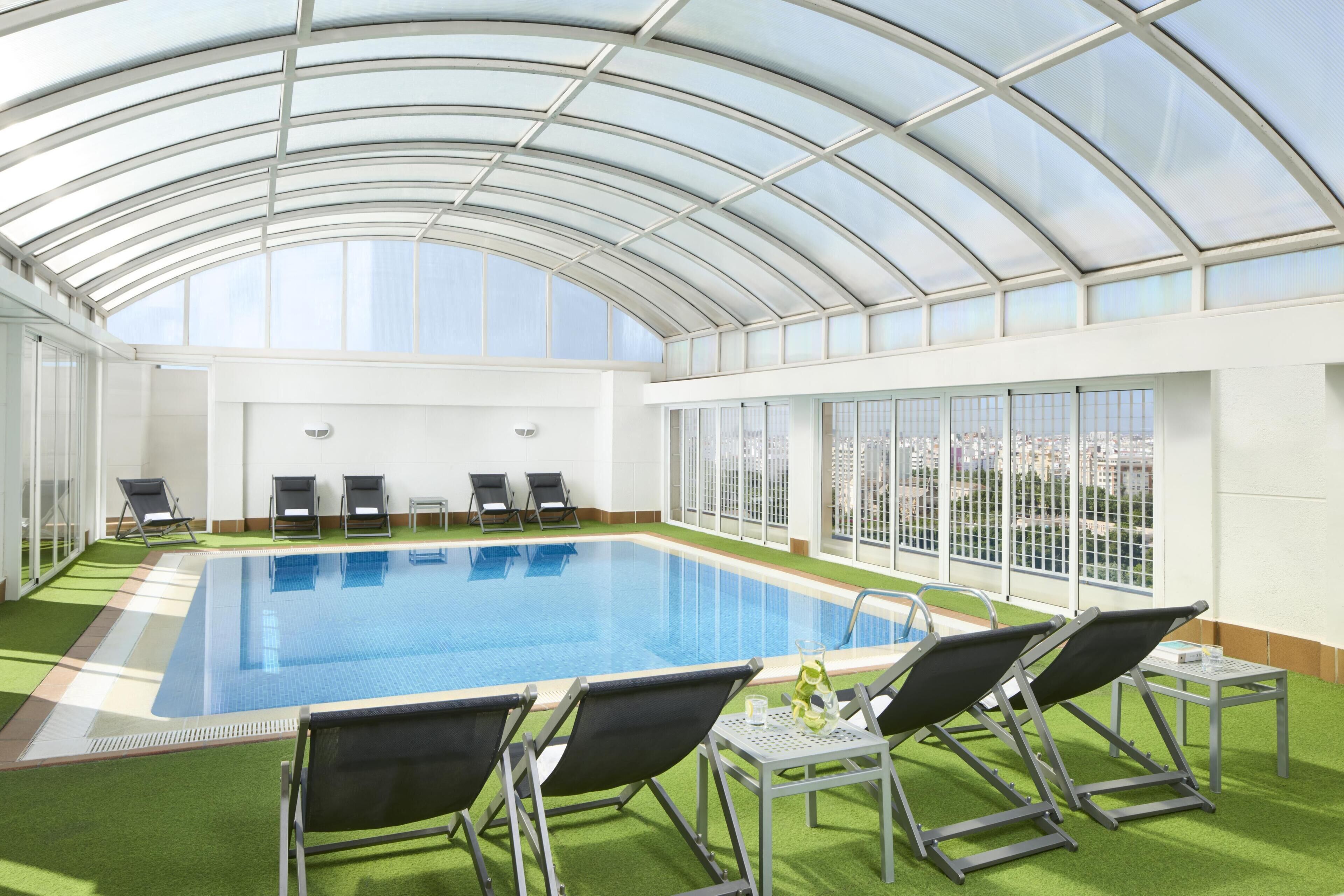 Piscina com teto de vidro, rodeada por espreguiçadeiras e vista para a cidade.