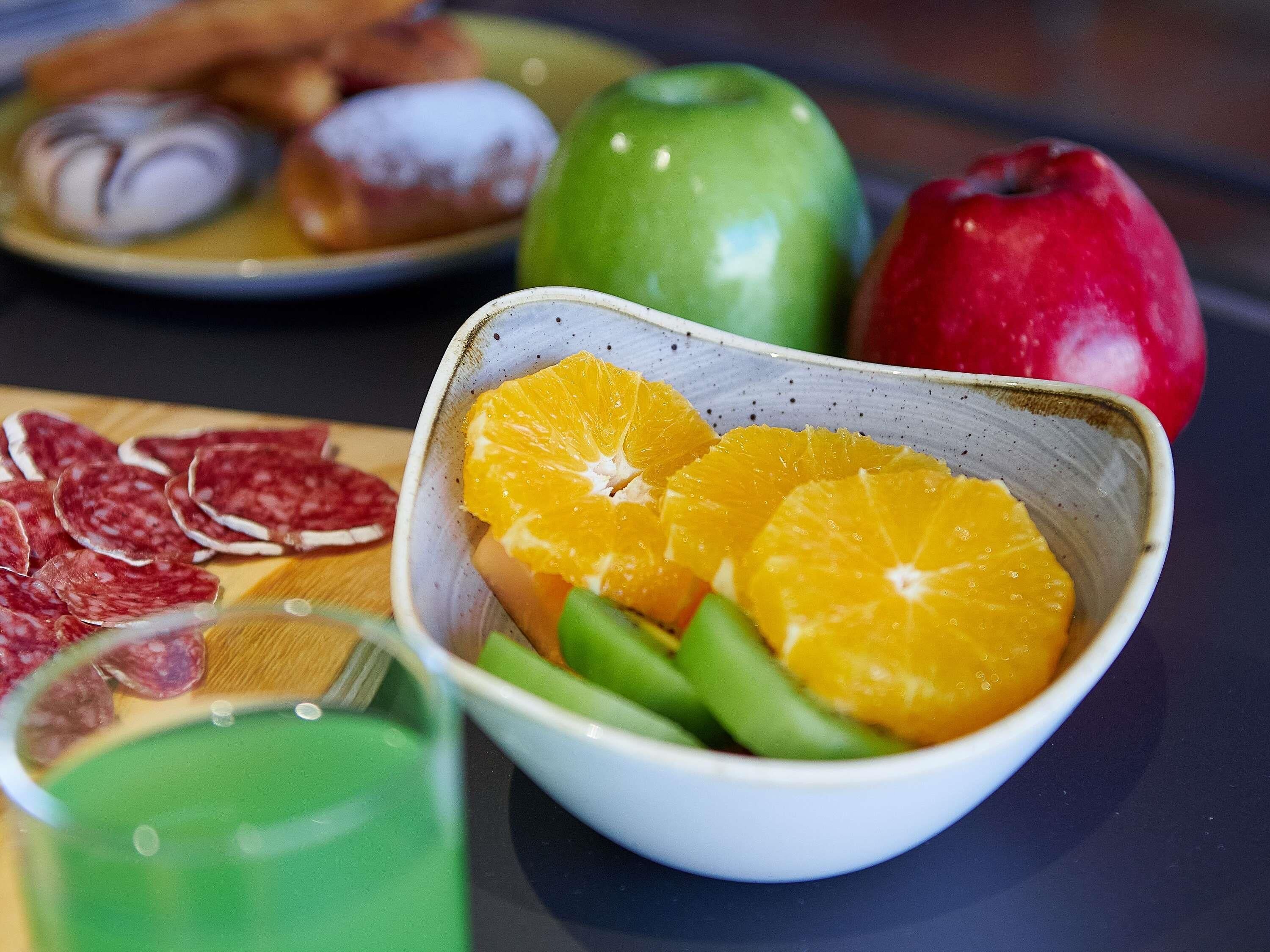 A bowl of sliced oranges and green beans, a plate with sliced salami, and a green apple on a table.
